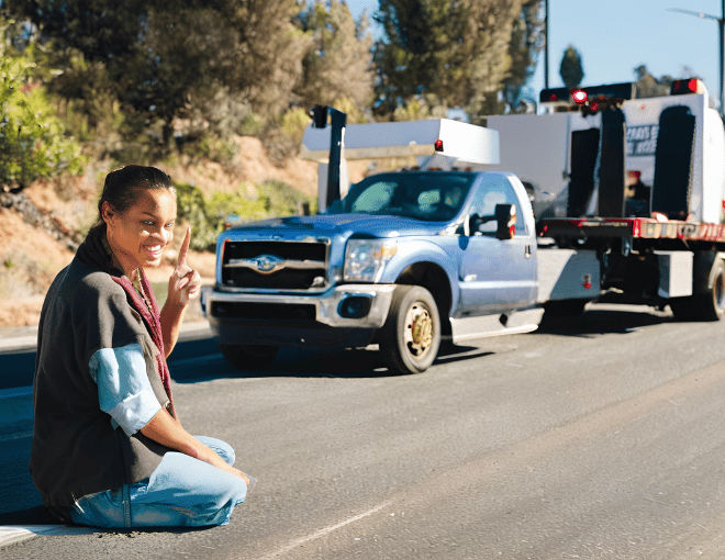 A hitchhiker stands by a roadside with a thumb out, a tow truck visible in the background, highlighting tips for dealing with towing in San Jose.